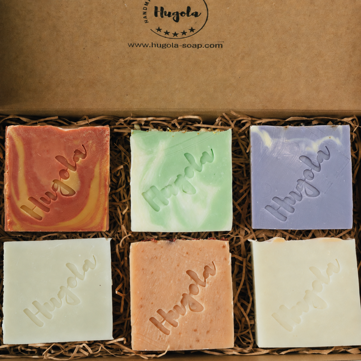 Six square bars of Hugola handmade soap in assorted colors arranged in a brown kraft gift box with shredded paper filler