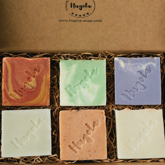 Six square bars of Hugola handmade soap in assorted colors arranged in a brown kraft gift box with shredded paper filler